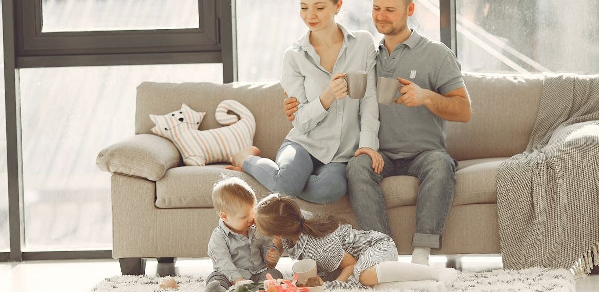 Cheerful family smiling while spending weekend together sitting parents sitting on comfortable sofa while children resting on carpet and playing