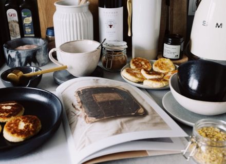 One portion of homemade cheese pancakes in frying pan and another on plate locating near opened stylish magazine among kitchen utensils and appliances in bright kitchen at home