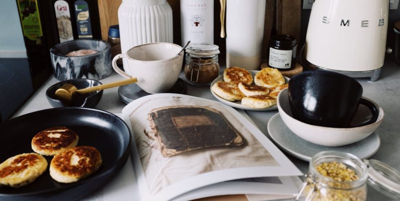 One portion of homemade cheese pancakes in frying pan and another on plate locating near opened stylish magazine among kitchen utensils and appliances in bright kitchen at home