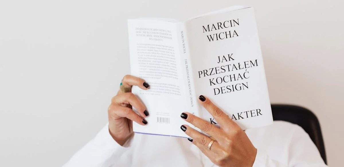 Anonymous female in white shirt with gold chain on neck reading interesting book covering face while sitting on black chair