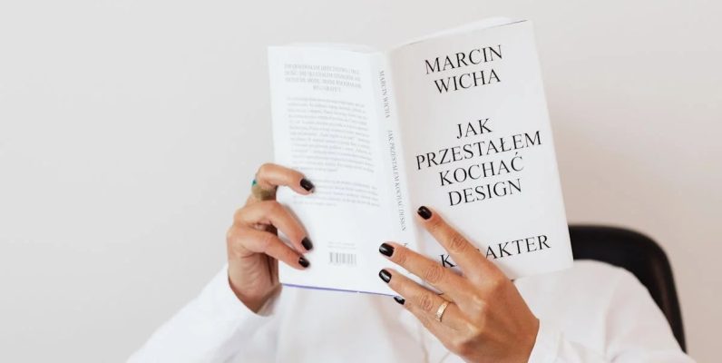 Anonymous female in white shirt with gold chain on neck reading interesting book covering face while sitting on black chair